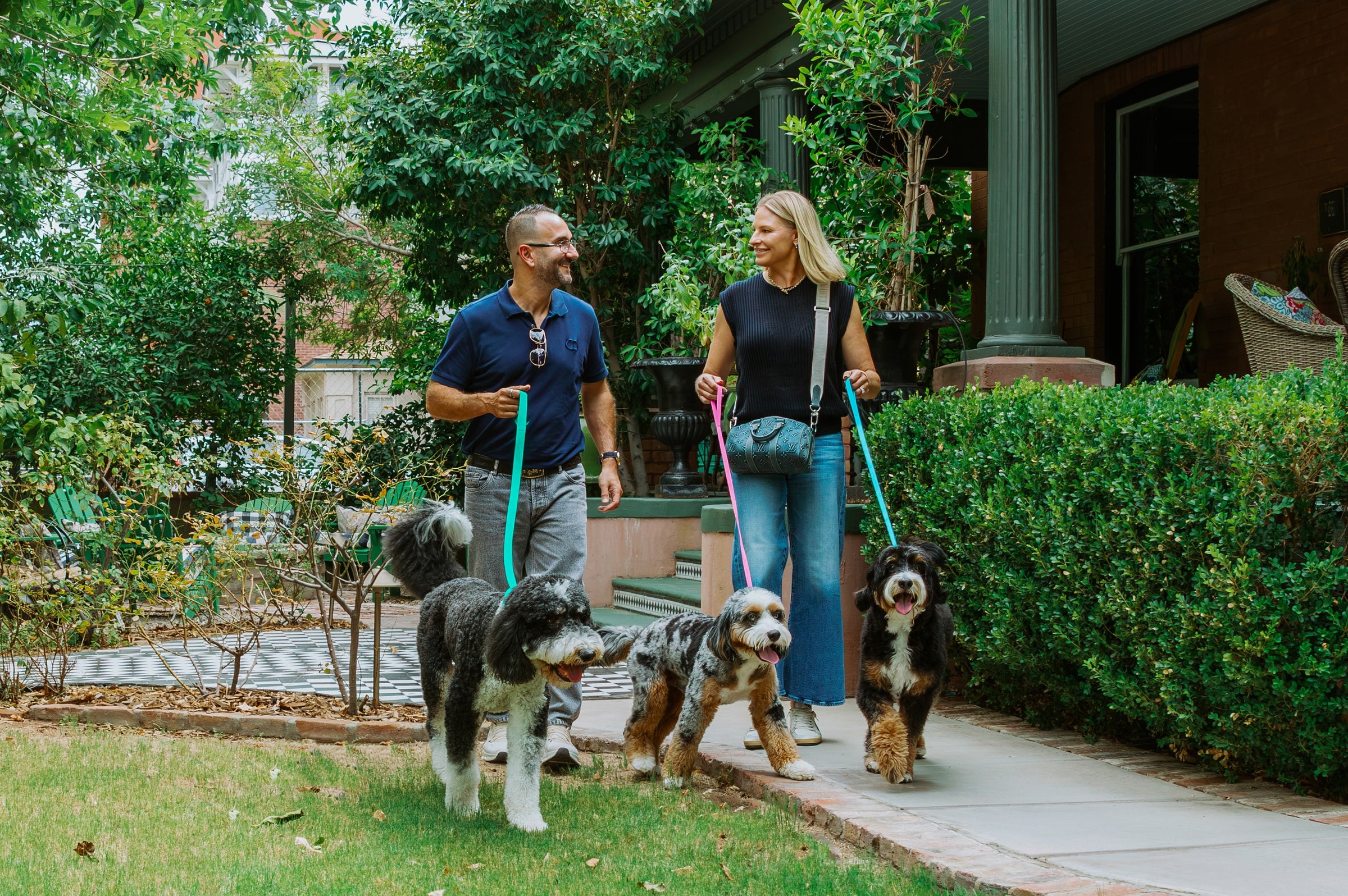 A man and a woman walking three dogs on colorful leashes along a paved path in a landscaped garden area with green hedges and a house in the background.