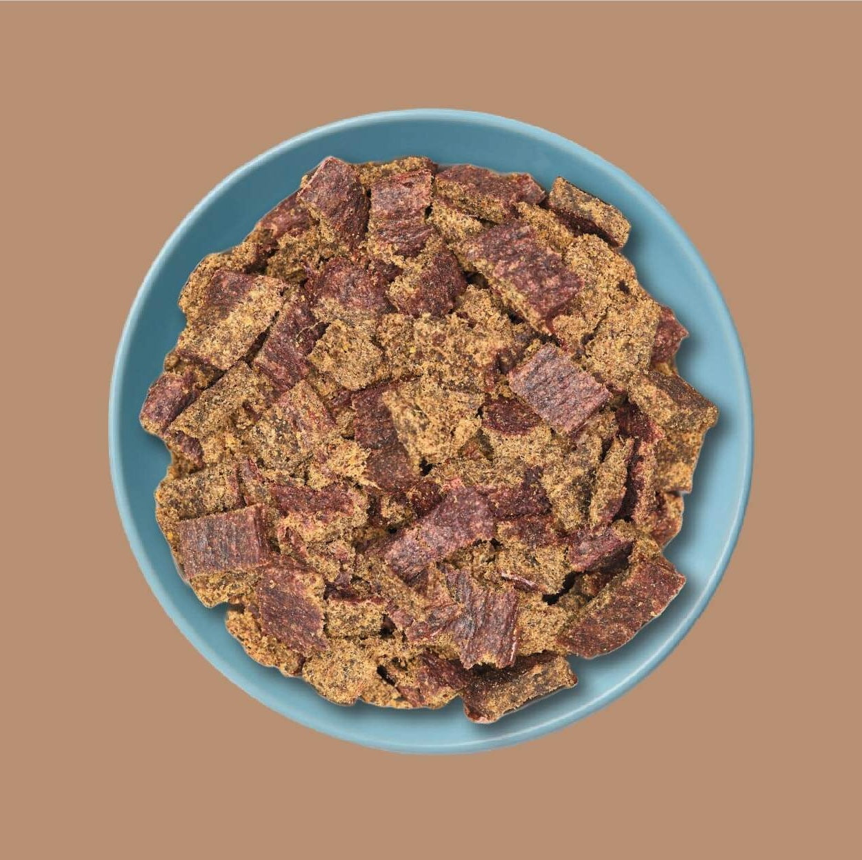 A top-down view of a pile of air-dried dog food, including both plain brown pieces and marbled pieces with reddish-brown flecks, served on a light blue plate against a solid brown background.