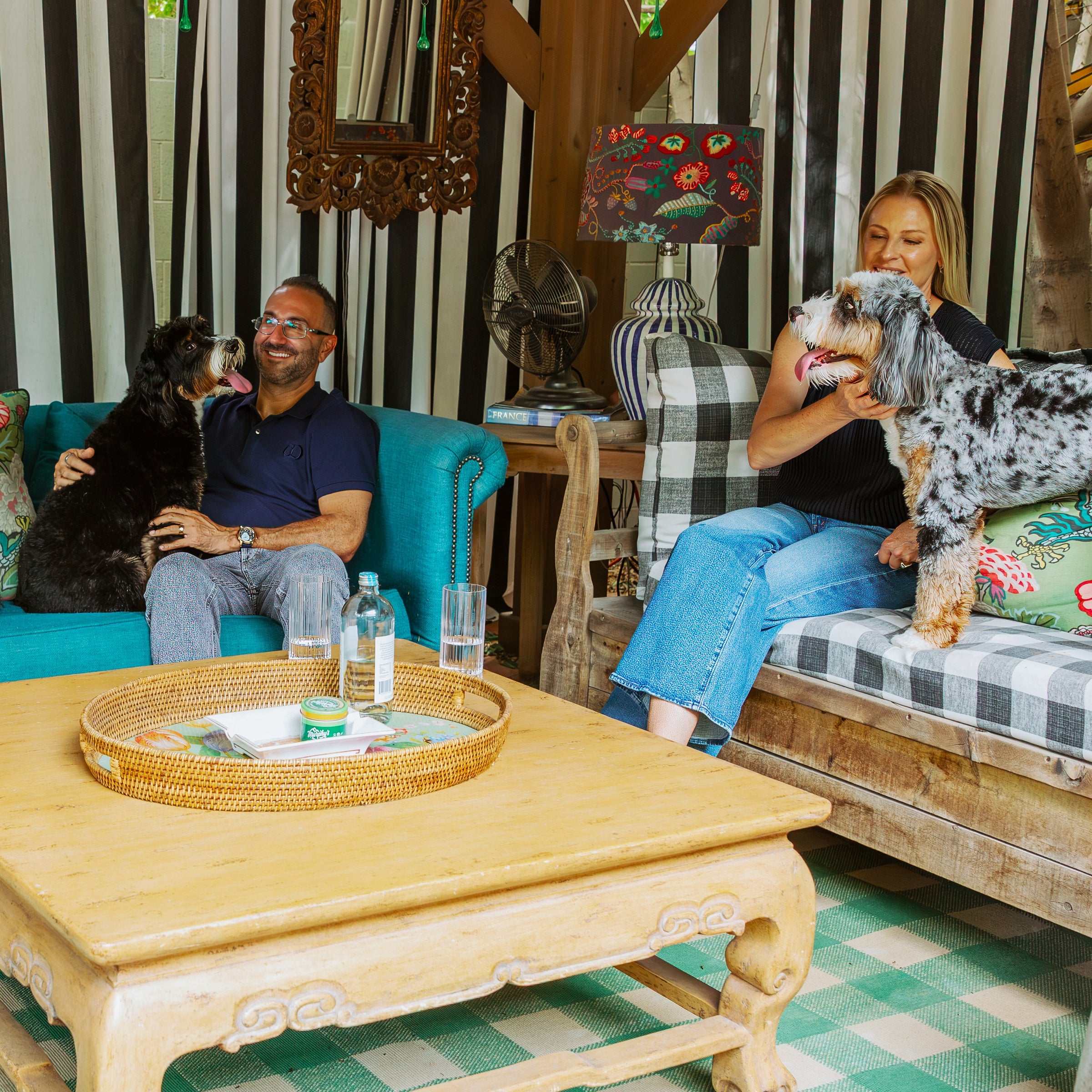 A cozy indoor living room scene with Unconditional founder and his wife sitting on sofas with dogs. A woman in a black top and blue jeans holds a black-and-white dog on a rustic wooden bench, while a man in a navy shirt sits on a turquoise sofa holding a black dog.